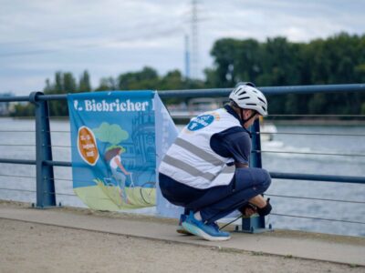 Person mit Fahrradhelm befestigt ein Banner an einem Geländer am Rheinufer
