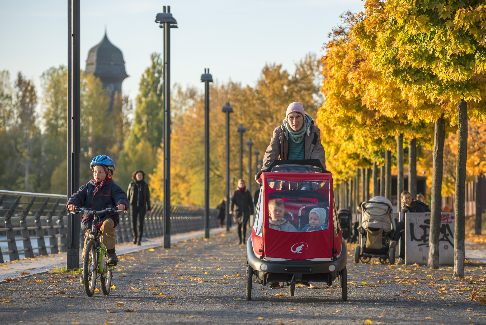Radfahrerin auf einem Lastenrad mit 2 Kindern. Daneben ein kleines Kind auf einem Fahrrad. Im Hintergrund sind Bäume im Herbst zu sehen